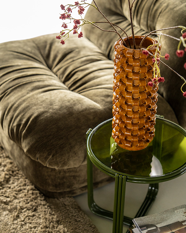 Groene velvet fauteuil met groene ronde bijzettafel met glazen blad en mustard keramieken vaas erop.